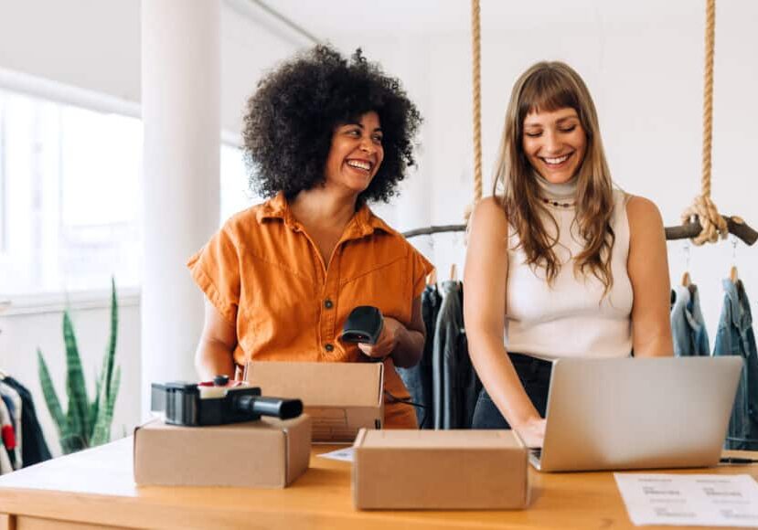 Cheerful clothing store owners preparing online orders for shipping Cheerful clothing store owners preparing online orders for shipping. Two happy businesswomen smiling at business tax while working together in a thrift store. Female entrepreneurs running an e-commerce small business.